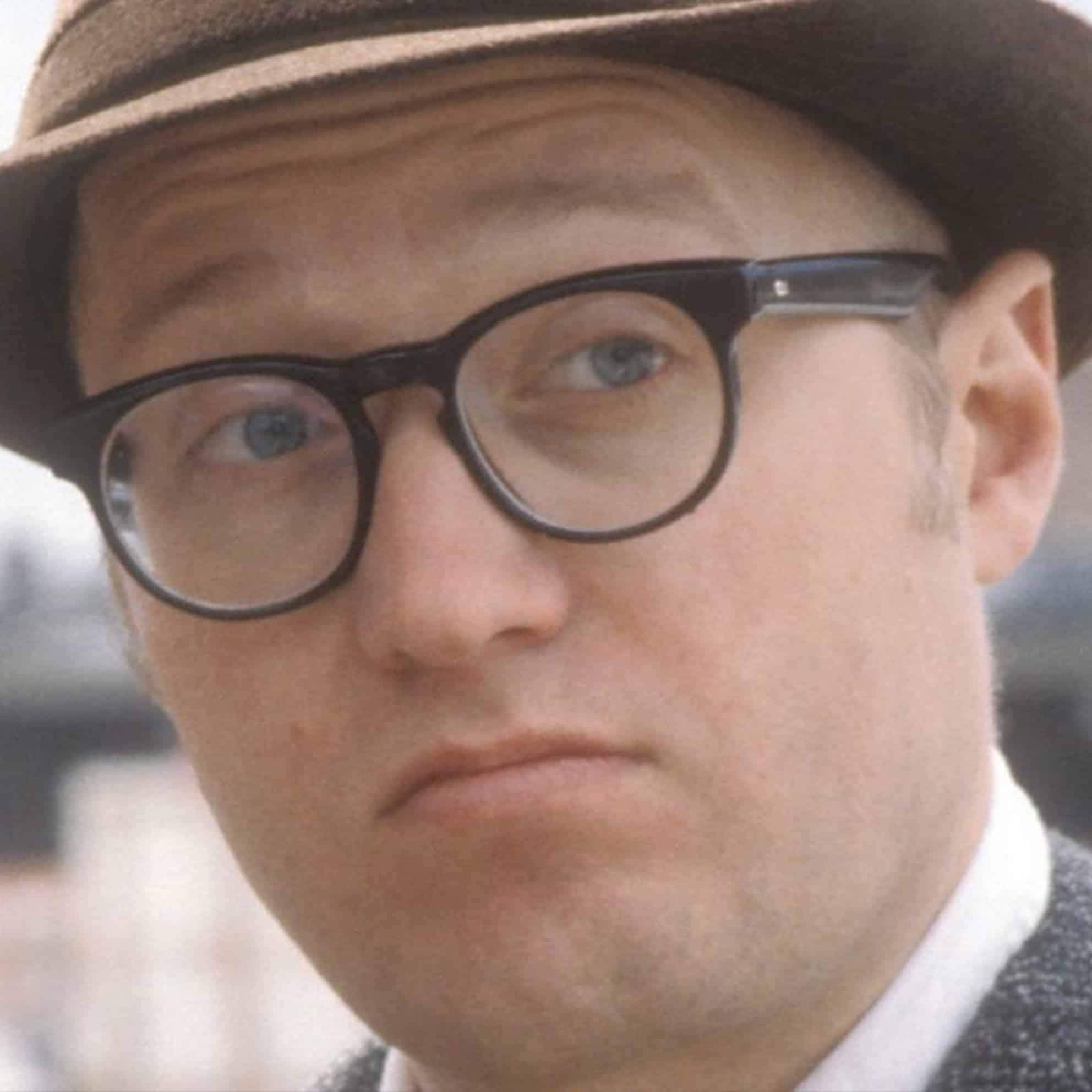 A member of The Print 4 Team, wearing glasses and a hat, gazes slightly to the side with an inquisitive expression.